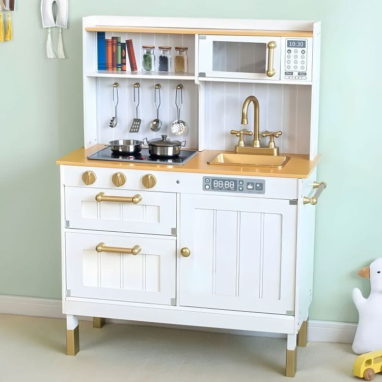2. White play kitchen with gold handles, featuring a sink, stove, and hanging utensils, set against a light green wall.