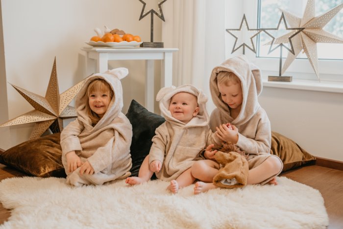 1. Three children wearing RÄTT's Long-Sleeve Poncho TEDDY in beige, sitting on a fluffy rug in a cozy room setting