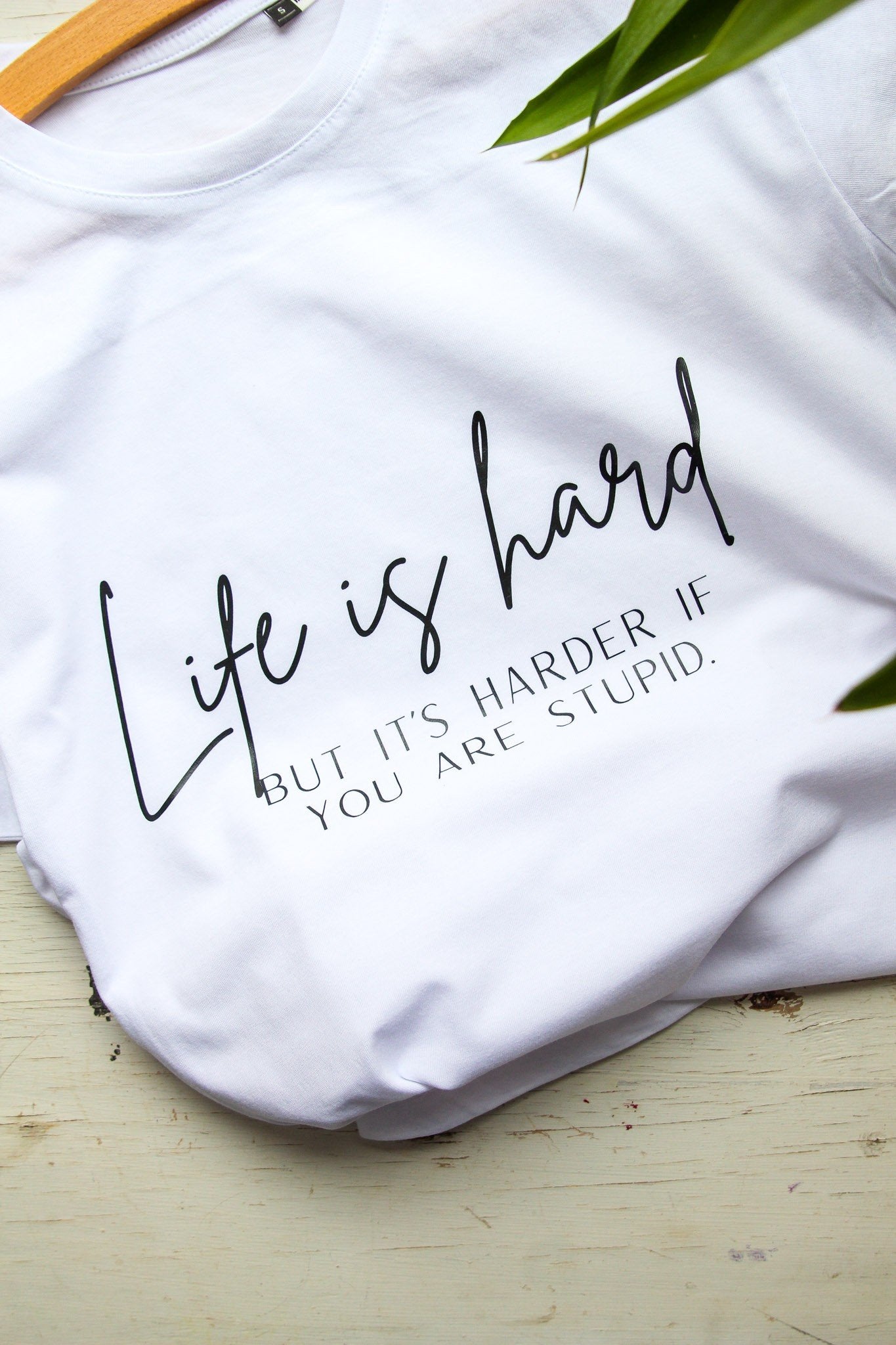 1. White oversized t-shirt with 'Life is hard but it's harder if you are stupid' text, laid on wooden surface with green plant accent