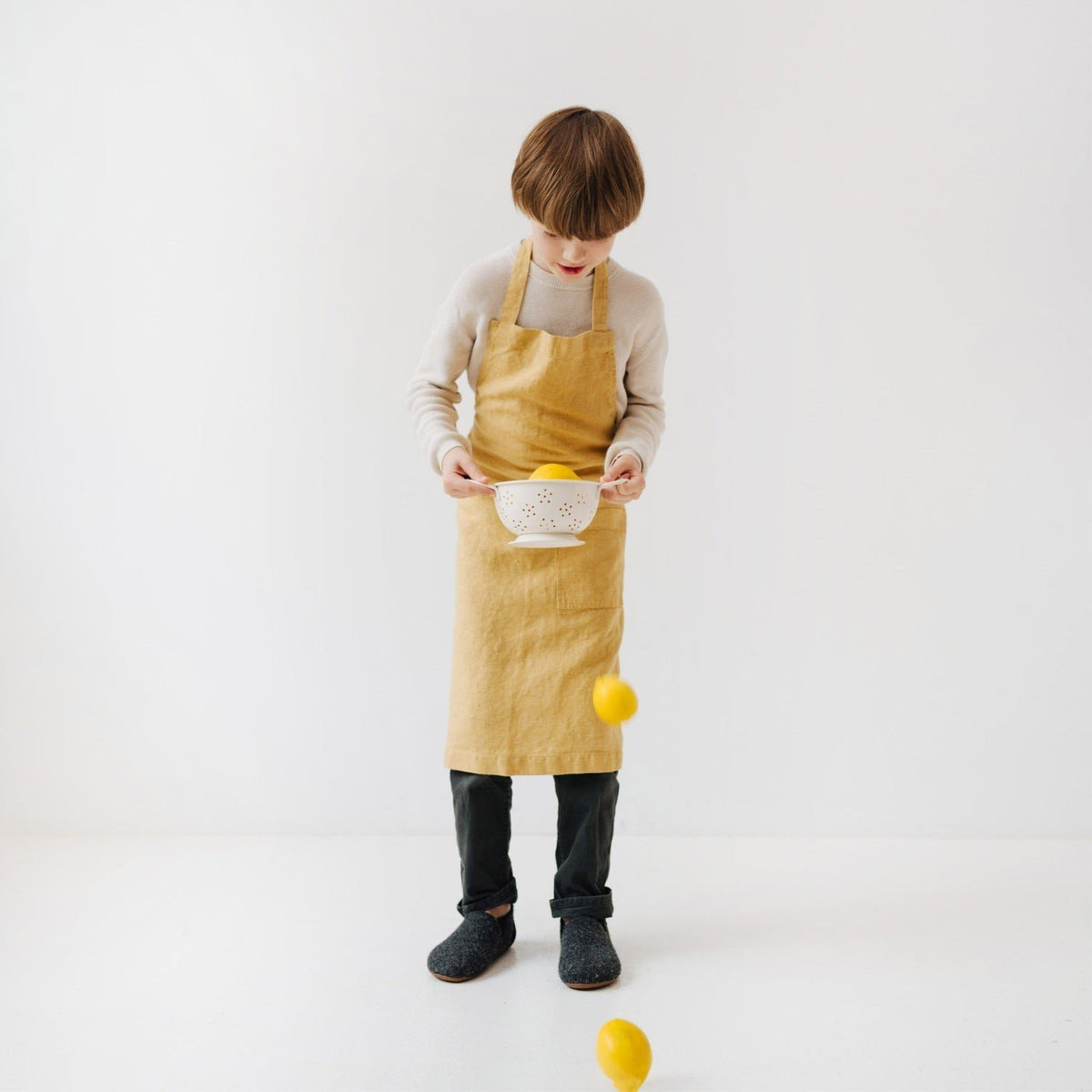 2. Child in honey-colored linen apron looking down at falling lemons in a white room