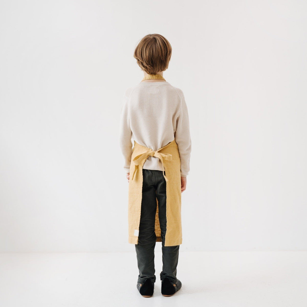 4. Back view of child wearing honey-colored linen apron with tied closure in a white room
