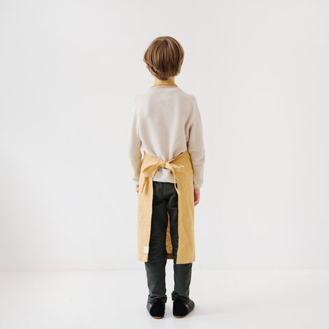 4. Back view of child wearing honey-colored linen apron with tied closure in a white room