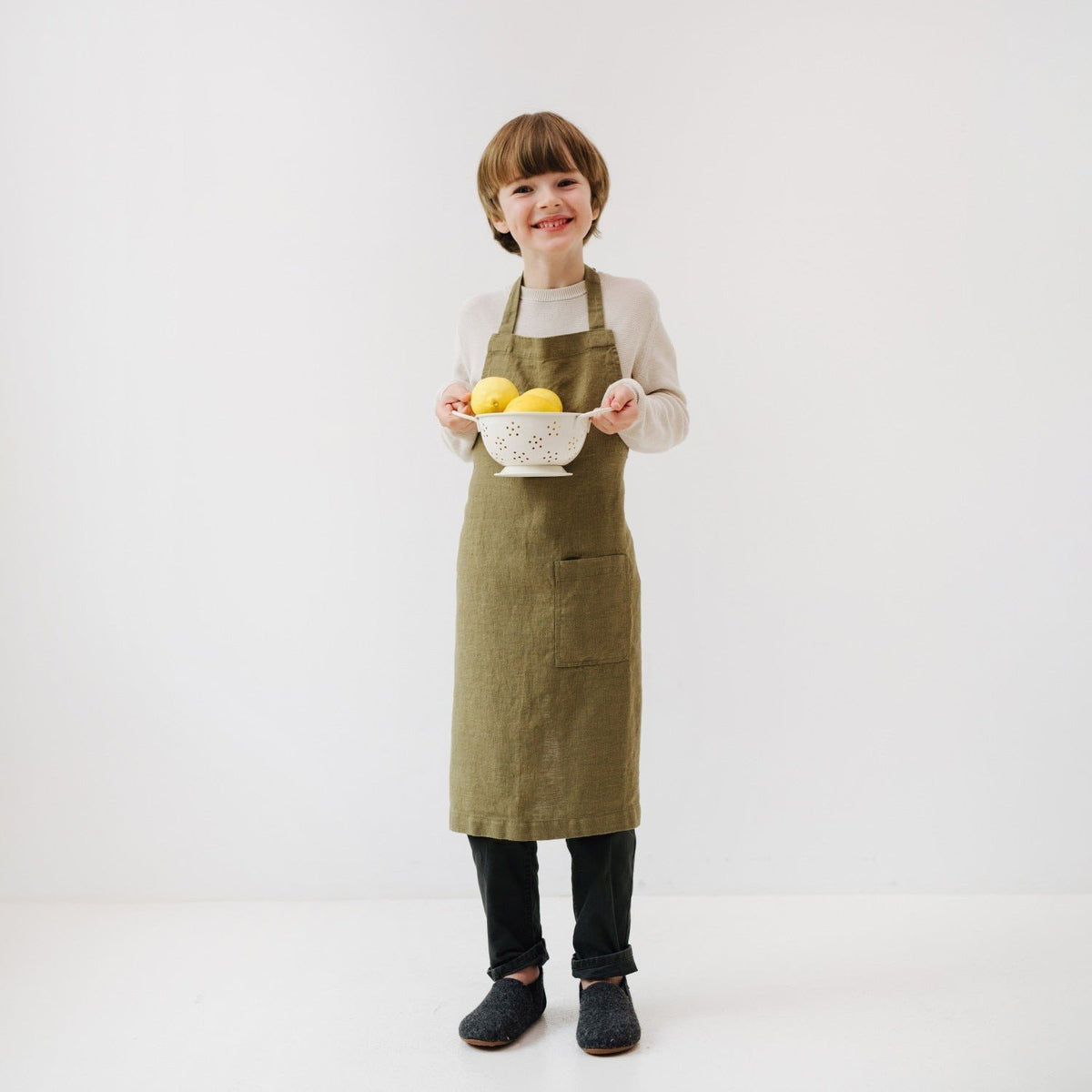 2. Child in olive green linen apron holding a colander with lemons, smiling in a white room