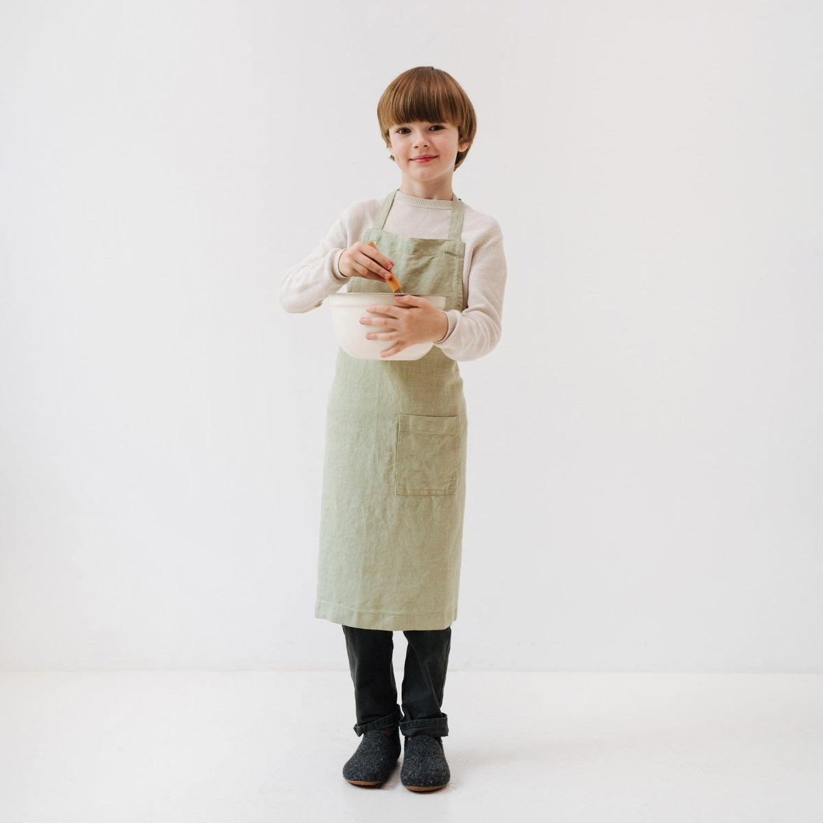 4. Child in sage green linen apron holding mixing bowl and spoon, standing in white studio