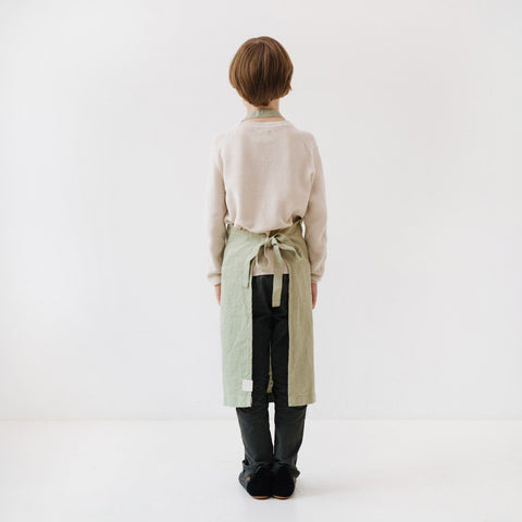 5. Back view of child wearing sage green linen apron with adjustable tie closure, standing in white studio