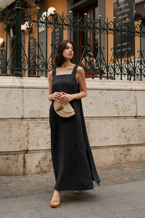 1. Woman wearing black linen Fugue dress with wide straps and square neck, holding a woven clutch, standing on a cobblestone street with iron fence background