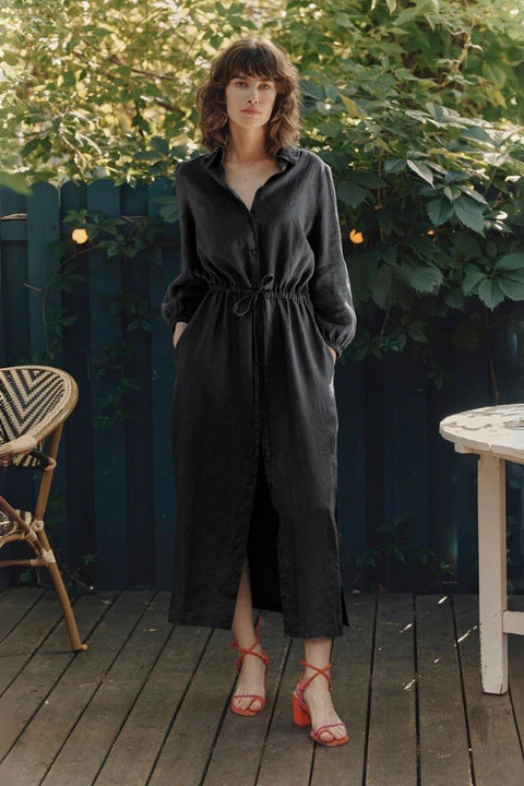 1. Woman wearing black long linen shirt dress with side slits and adjustable drawstring belt, standing on a wooden deck with greenery in the background