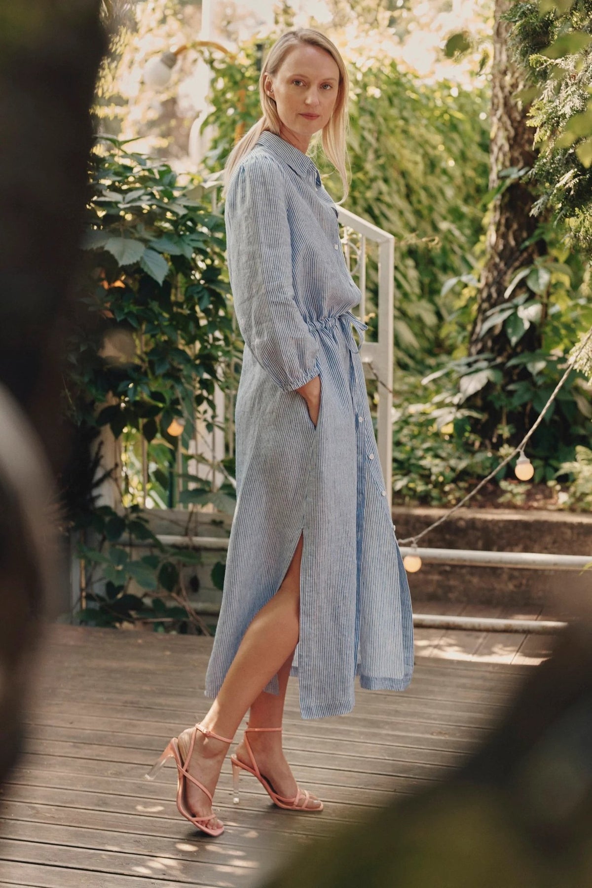3. Side view of woman in blue and white striped linen dress with button closure and side slits, standing on wooden deck