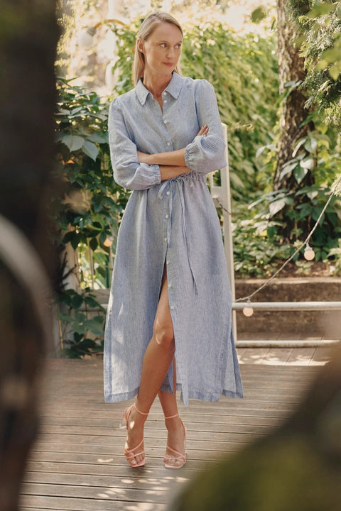 1. Woman wearing blue and white striped linen dress with side slits and adjustable belt, standing on a wooden deck surrounded by greenery