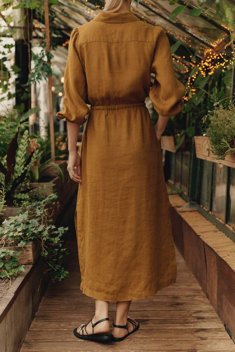 3. Back view of woman in bronze linen Rhapsody dress with elasticated waist, in a greenhouse