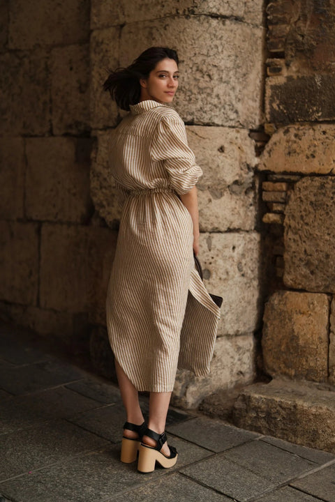 3. Back view of woman in bronze and white striped linen dress with elasticated cuffs and side slits, standing by a stone wall
