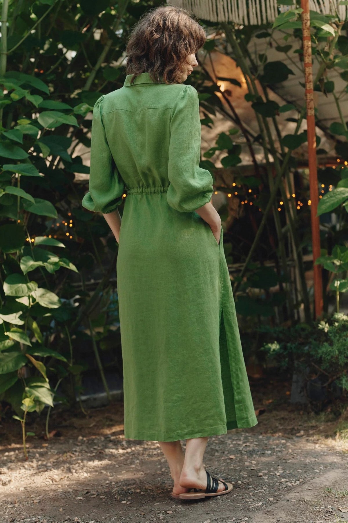 2. Back view of green linen long shirt dress with elasticated cuffs and side slits, worn by woman in garden