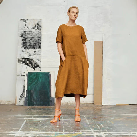 1. Woman wearing bronze linen A-line dress with short sleeves and round neck, standing in an art studio setting with abstract paintings in the background