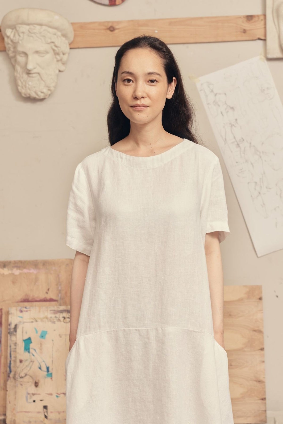 2. Woman in optical white linen dress with round neck and front pockets, standing in art studio