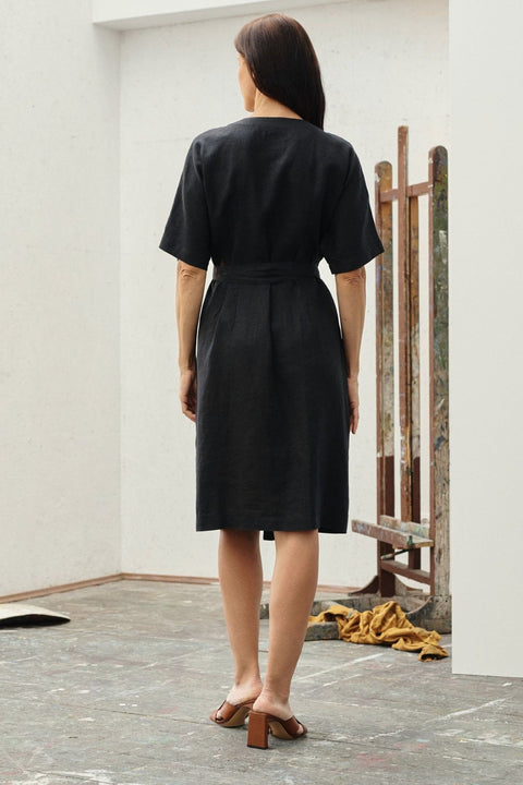 2. Back view of black linen wrap dress with short sleeves and tied waist, worn by a woman in an art studio