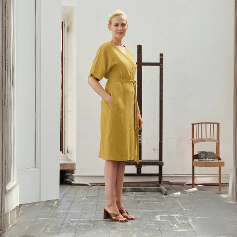 1. Woman wearing lemon curry linen wrap dress with V-neck and tied fastening, standing in a bright room with wooden floors and a chair in the background