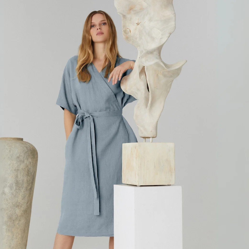 1. Woman wearing blue linen wrap dress with tie fastening, standing next to abstract sculpture in studio setting
