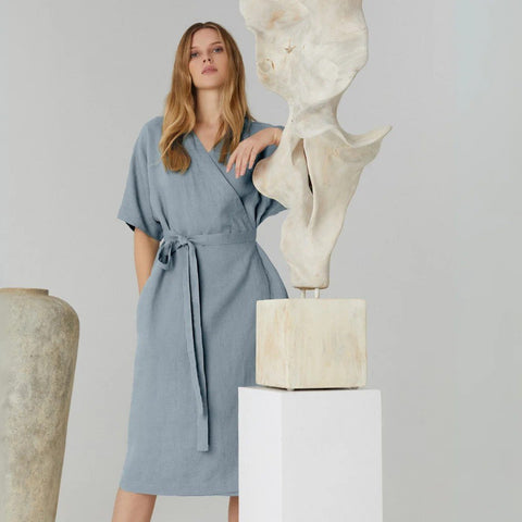 1. Woman wearing blue linen wrap dress with tie fastening, standing next to abstract sculpture in studio setting