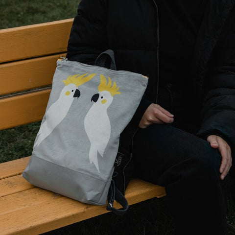 2. Muni Cockatoo backpack in grey velvet with cockatoo design, placed on a bench next to a person in a black jacket