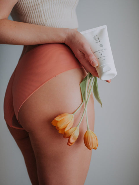 1. Woman holding LUUV Sensitive Intimate Wash and orange tulips against a neutral background, wearing peach underwear and a white top