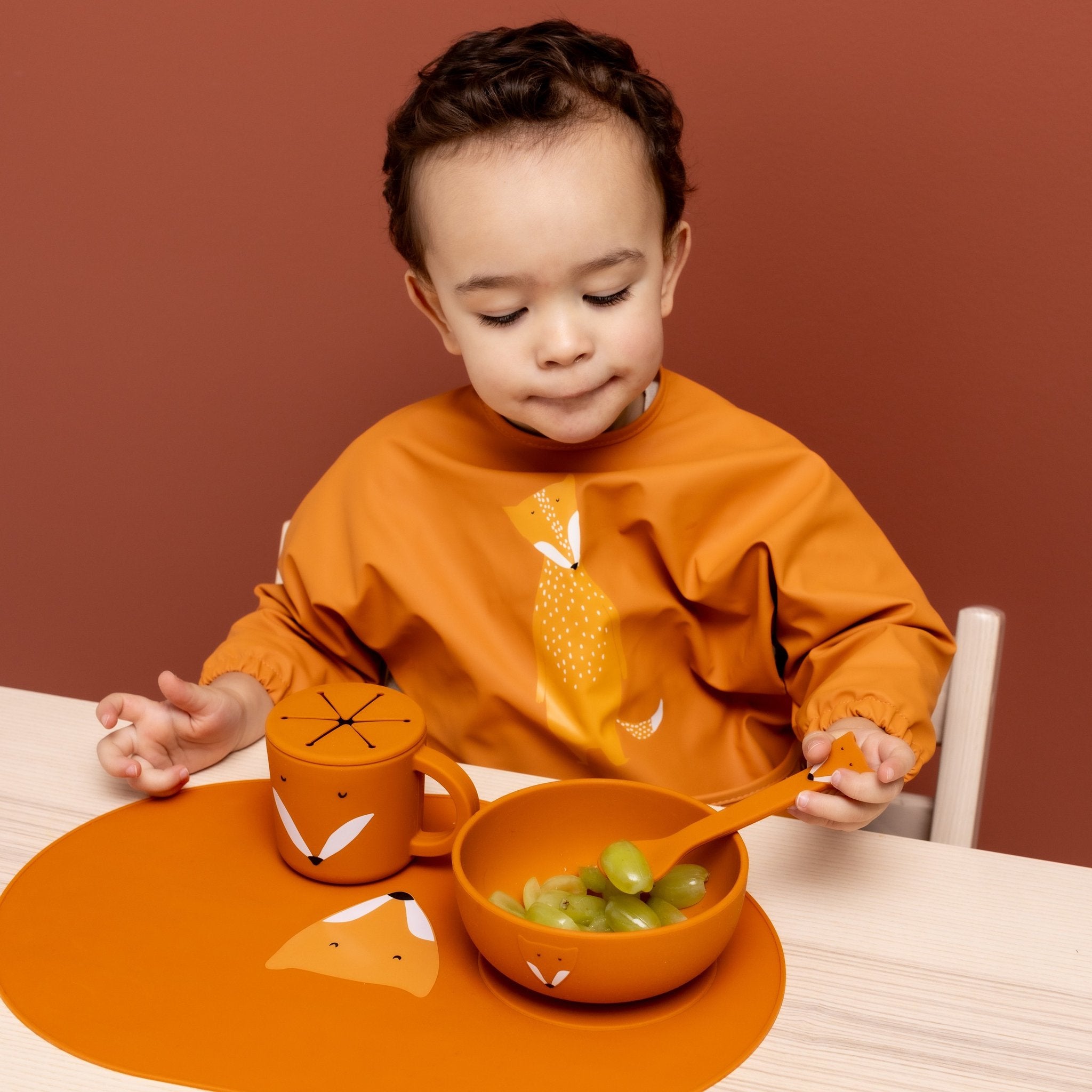 1. Child wearing orange Trixie Baby waterproof long sleeve bib with Mr. Fox design, sitting at table with matching fox-themed dinnerware