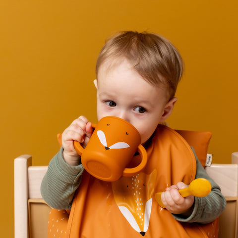1. Toddler using orange Mr. Fox silicone sippy cup with big handles, wearing matching bib, against mustard background