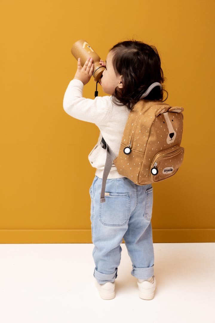 1. Child drinking from Trixie Baby Mr. Bear stainless steel bottle, wearing beige sweater and jeans, against mustard background