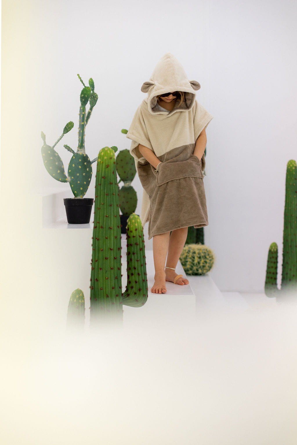 7. Child wearing beige Surfsheep kids bath poncho with hood and ears standing among cacti