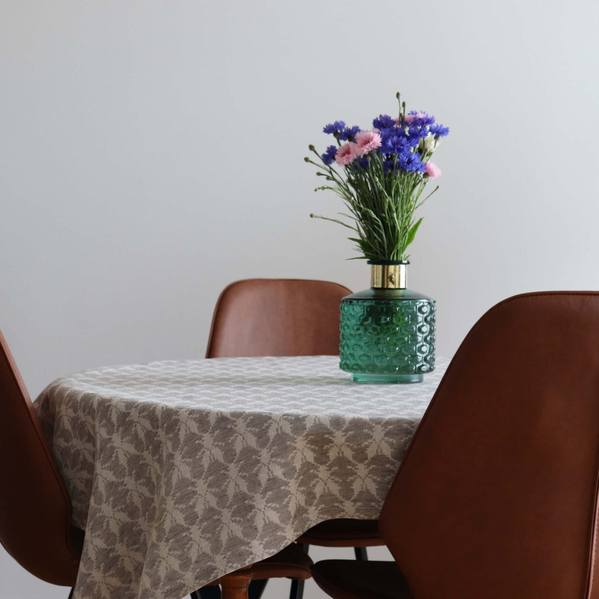 1. Dining table with LOKO Hanikatsi Fly tablecloth in beige linen-cotton blend, featuring Hiiumaa mitten pattern, set with brown chairs and a green vase with pink and blue flowers