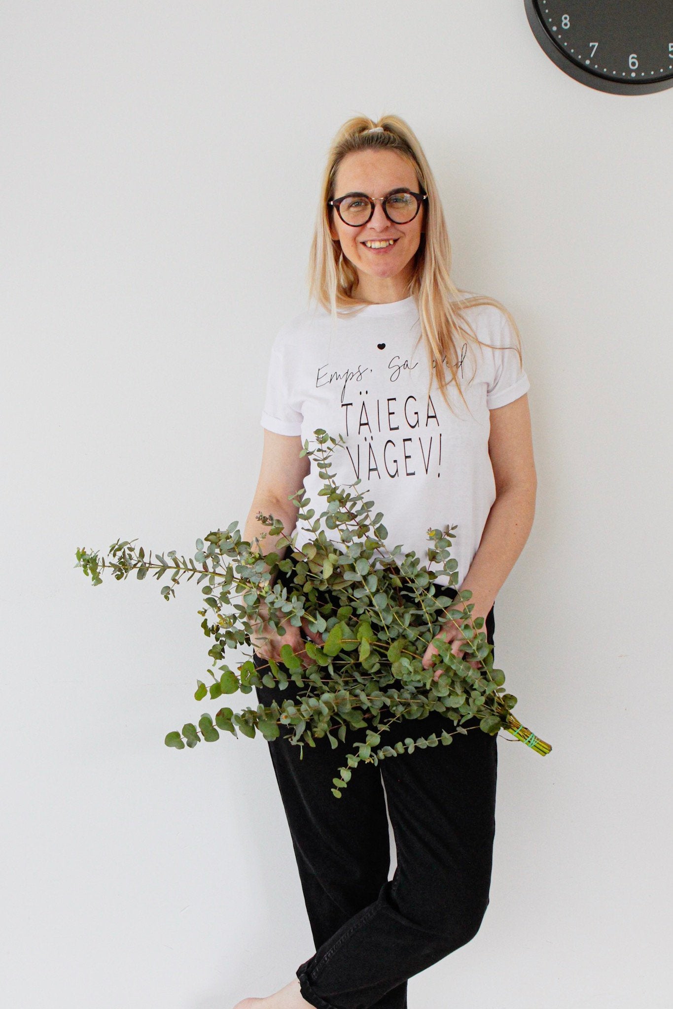1. Woman wearing Black Giraffe Brand white T-shirt with 'Emps, sa oled täiega vägev' text, holding eucalyptus branches