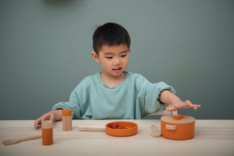 1. Young boy playing with Trixie Baby Wooden Kitchen Set, interacting with orange pot and utensils on table