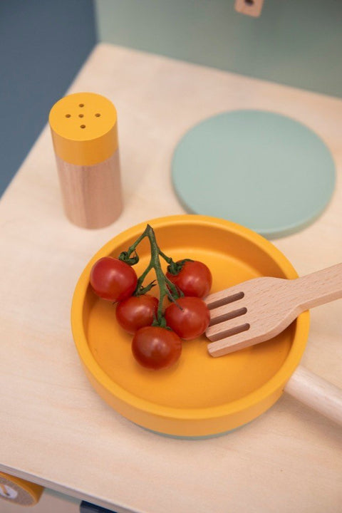 3. Close-up of Trixie Baby Mr. Lion wooden pan with cherry tomatoes and fork, next to a salt shaker on a play kitchen surface