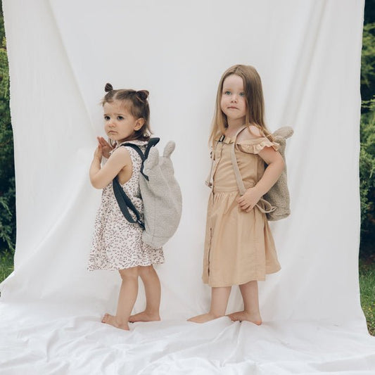 2. Two young girls lying on white fabric with Muni beige teddy backpacks, outdoor setting