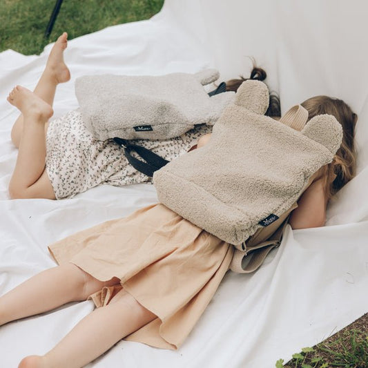 1. Two young girls wearing Muni beige teddy backpacks, standing on white fabric outdoors