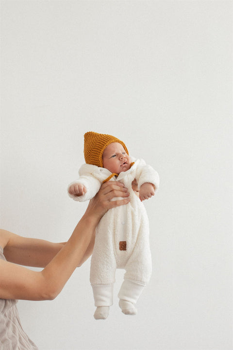 2. Baby held up wearing fuzzy ecru jumpsuit with bear ears hood and mustard knit hat