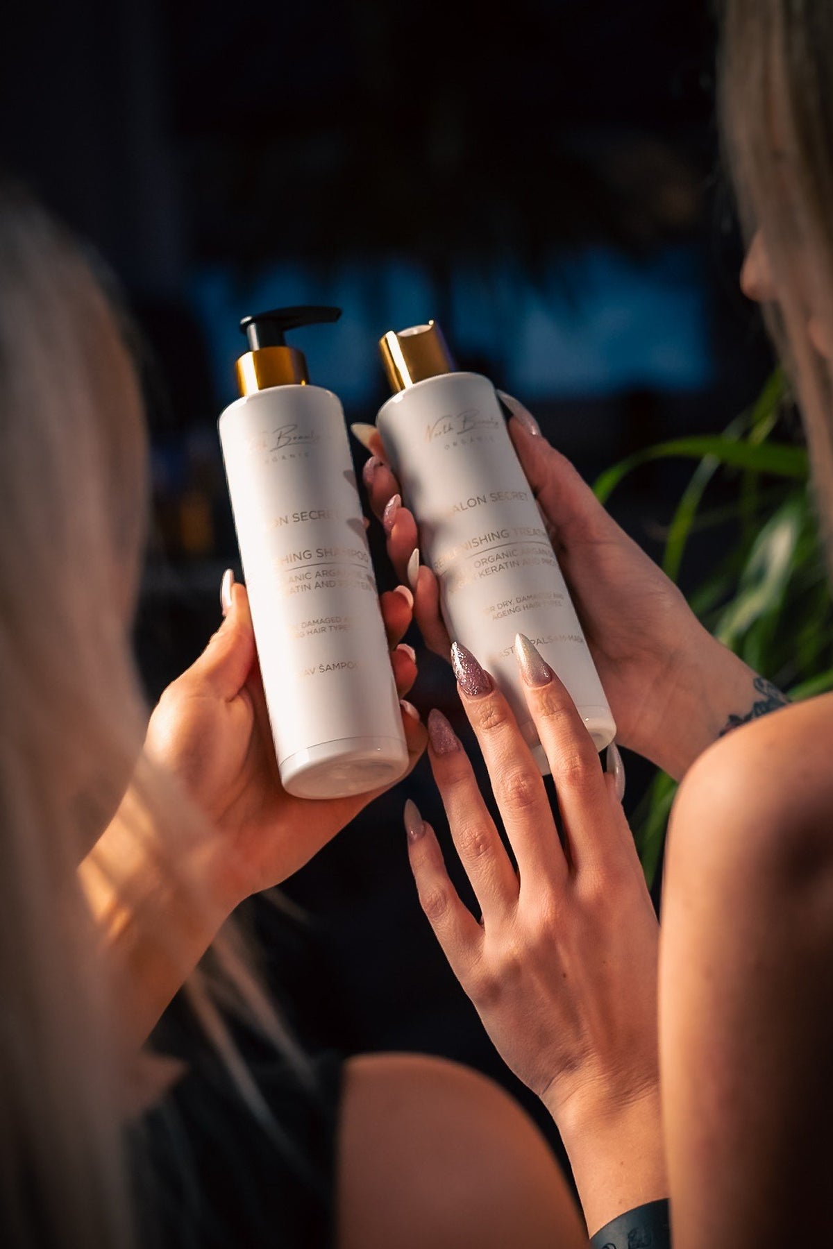 1. Two women holding North Beauty Salon Secret Replenishing Shampoo and Treatment bottles with gold caps in dimly lit room