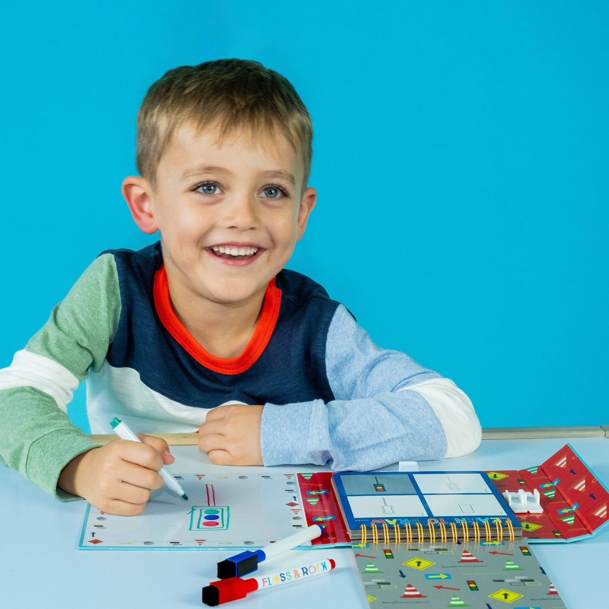 1. Smiling boy using Floss & Rock drawing kit with colorful markers and drawing pad on a blue background
