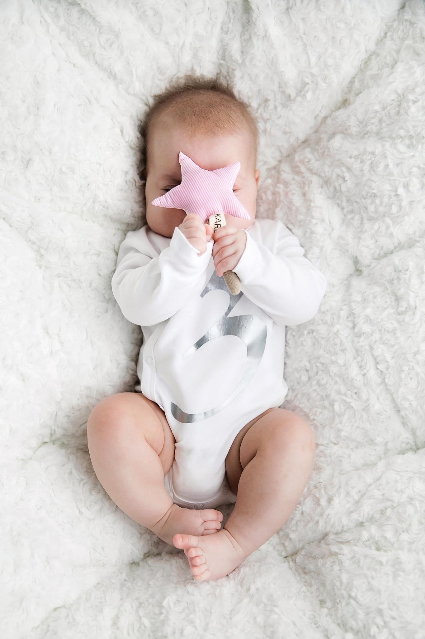 5. Baby in white onesie holding pink star-shaped Karloova Magic Wand on fluffy blanket