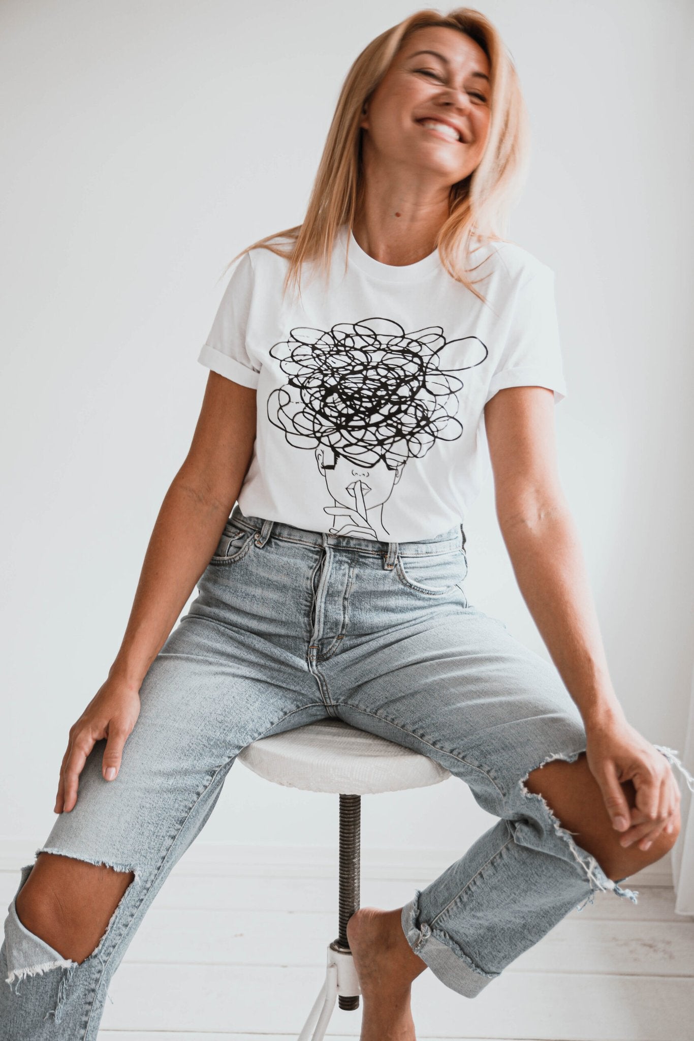 2. Women's white Šššhhh! T-shirt by Black Giraffe Brand with abstract face design, styled with ripped jeans in studio setting