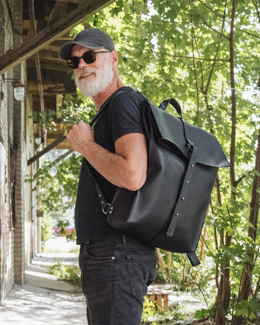 1. Man wearing black leather knapsack over shoulder, standing outdoors with trees in background