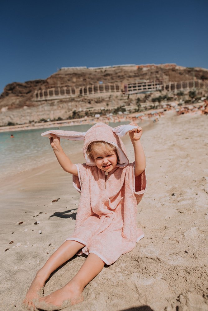 4. Child in pink RÄTT short-sleeve poncho sitting on the beach, holding hood ears
