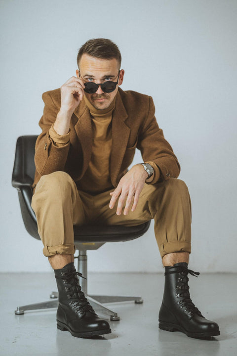 1. Man in brown jacket and tan pants wearing black M77 Norwegian combat boots, sitting on chair indoors, adjusting sunglasses