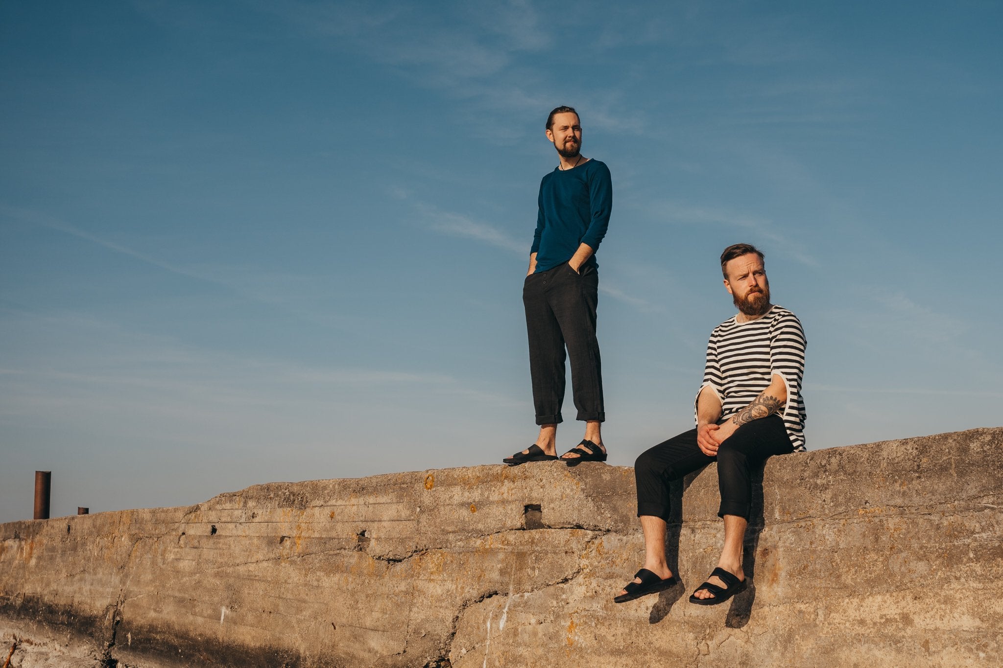 4. Two men wearing Madise Sandals in black, sitting and standing on a concrete ledge under a clear sky