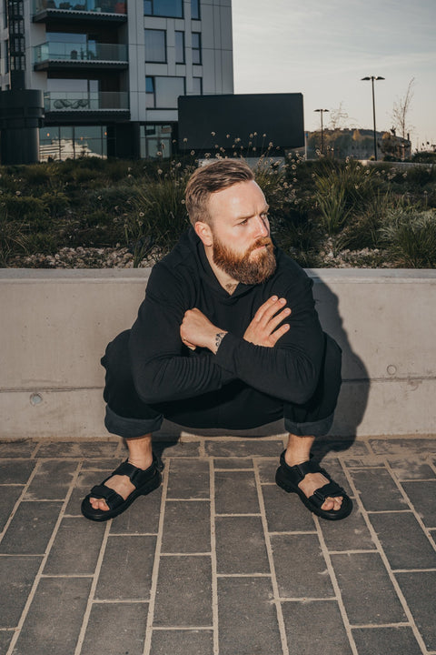 2. Man squatting outdoors wearing Omaking Lauri Leather Sandals in black, highlighting adjustable velcro straps