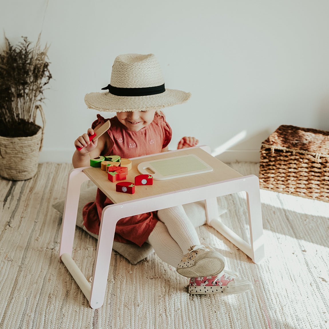 1. Girl in pink dress and straw hat sitting at small wooden desk with toy food in bright room