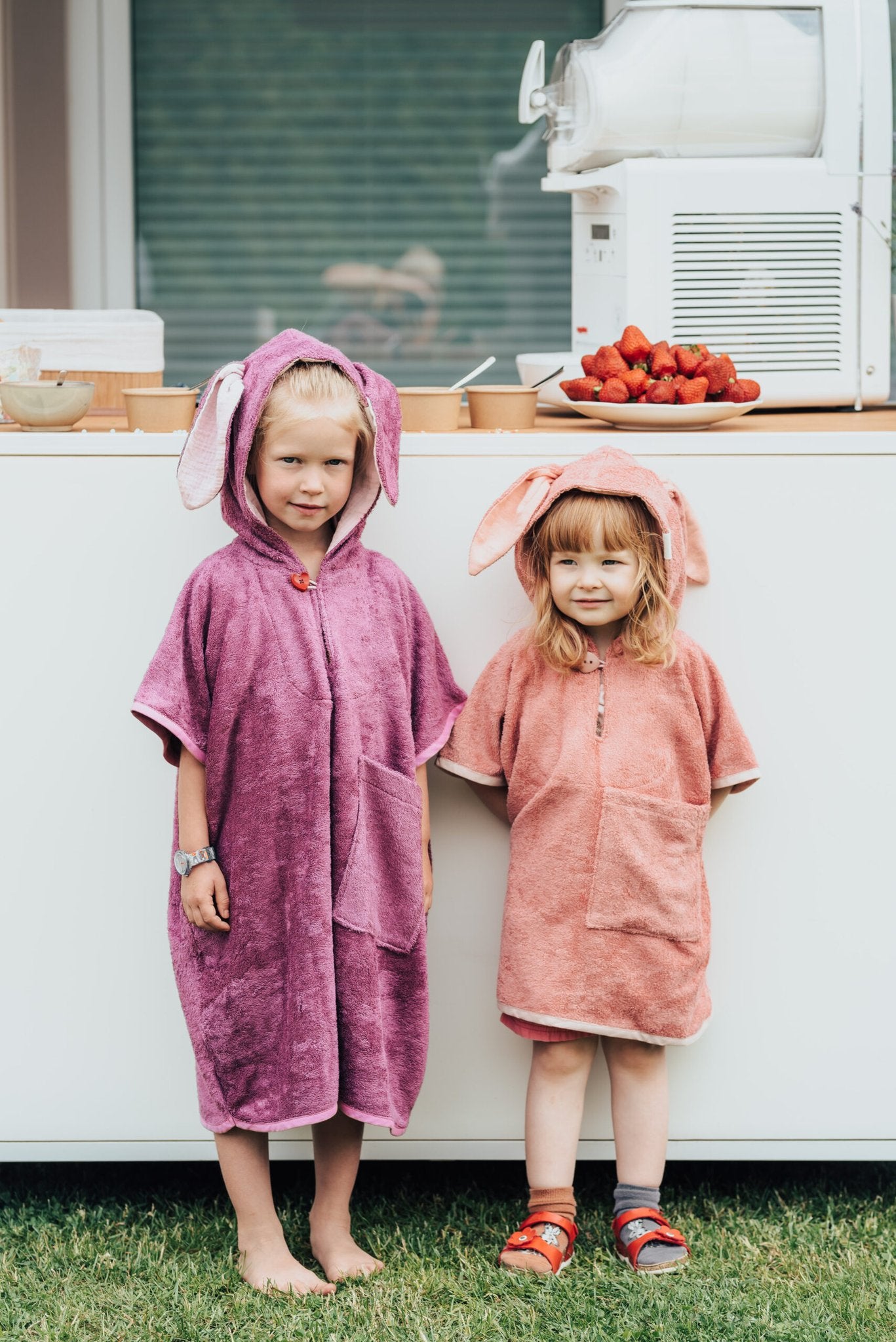 6. Two toddlers in RÄTT short-sleeve ponchos, purple and pink, standing by a table with strawberries