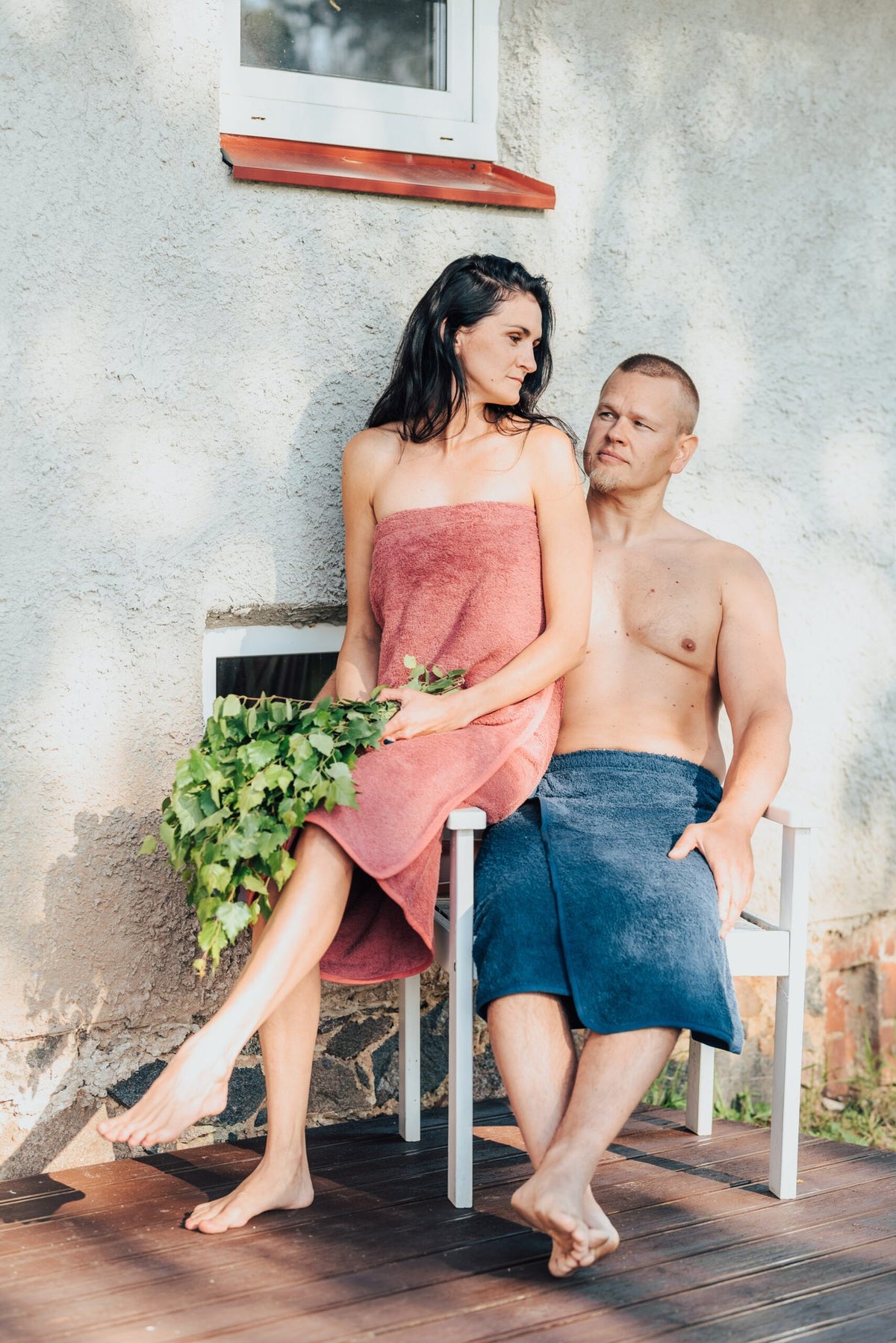 2. Woman in RÄTT dark dusty pink spa wrap and man in blue towel, sitting outdoors