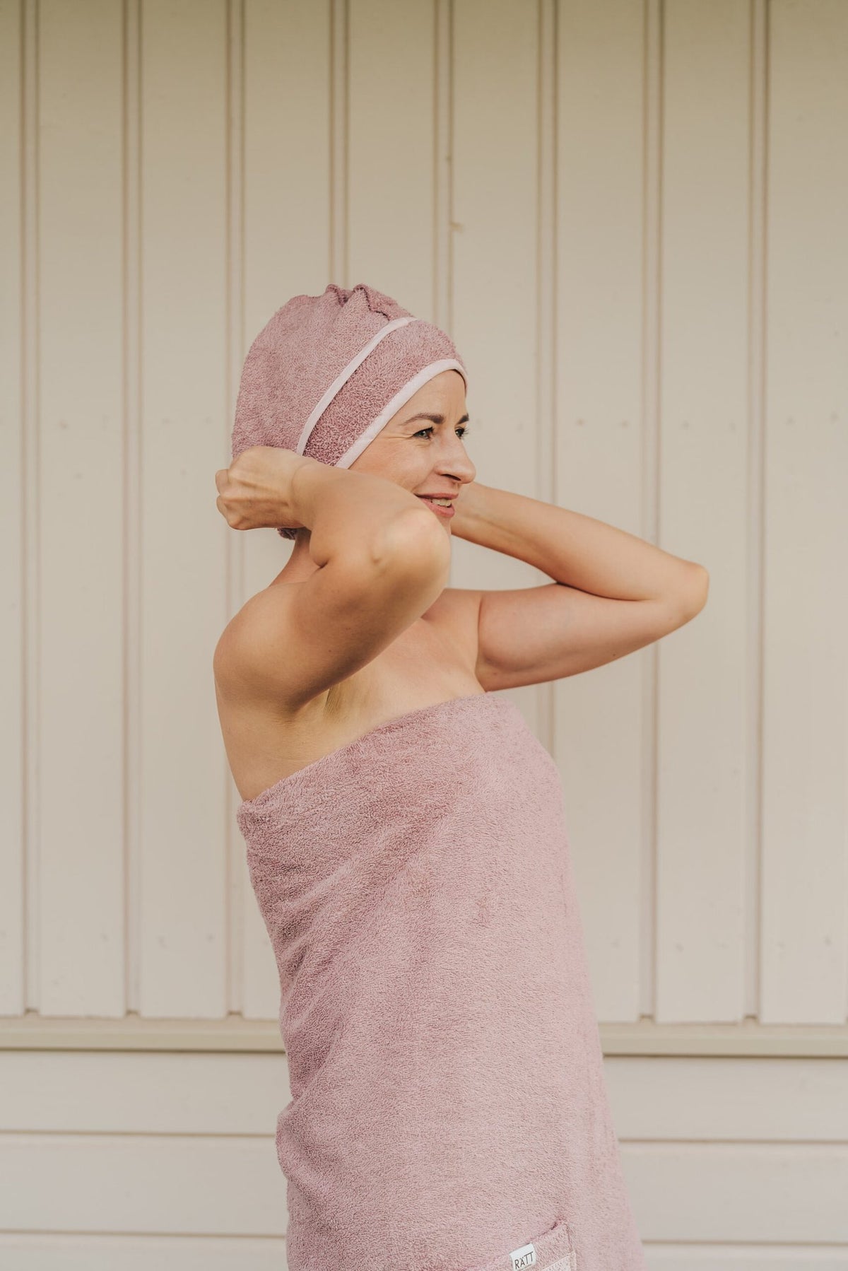 1. Woman wearing RÄTT cotton terry hair wrap in pink, adjusting it while wrapped in matching towel, lifestyle setting