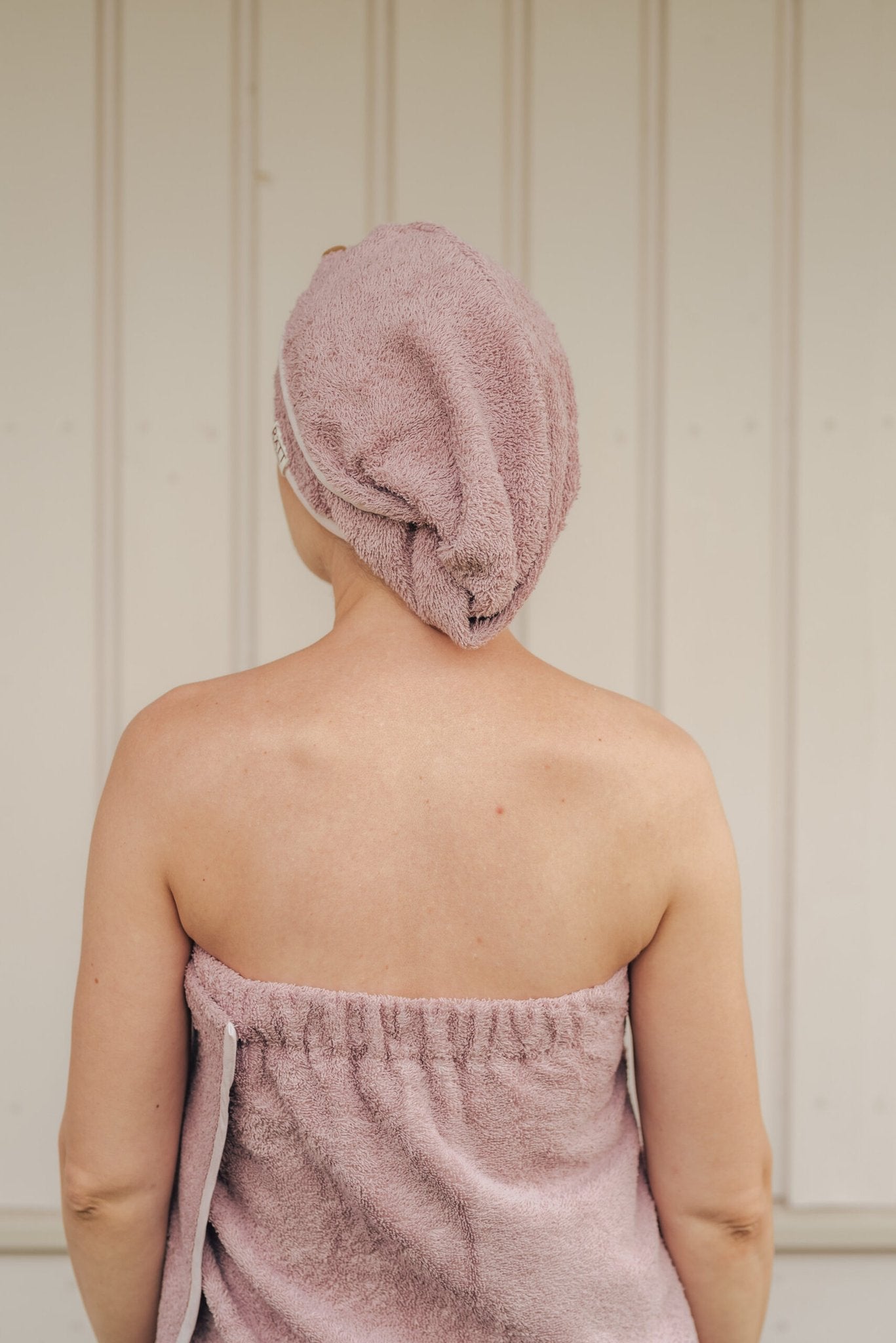 3. Back view of woman wearing RÄTT cotton terry hair wrap in pink, wrapped in matching towel, lifestyle setting