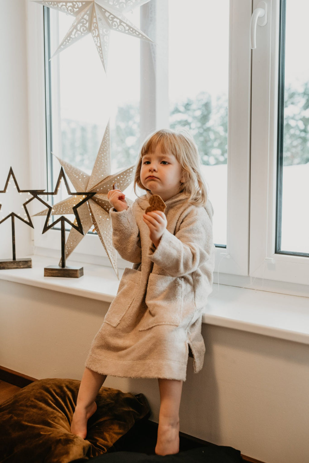 4. Child sitting on a windowsill wearing RÄTT's beige Long-Sleeve Poncho TEDDY, holding a cookie